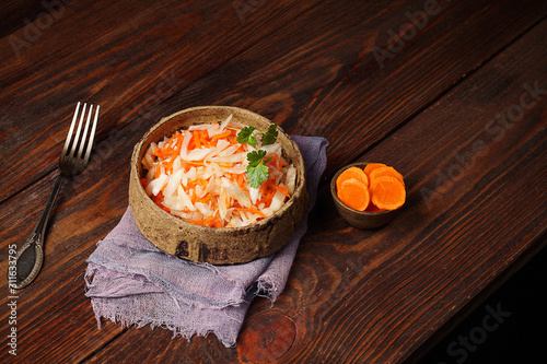 Fermented cabbage in a ceramic Cup with a sprig of parsley on a lilac napkin. Next to a fork, and a gravy boat with carrots cut into slices. Horizontal arrangement, wooden background, copy space. photo