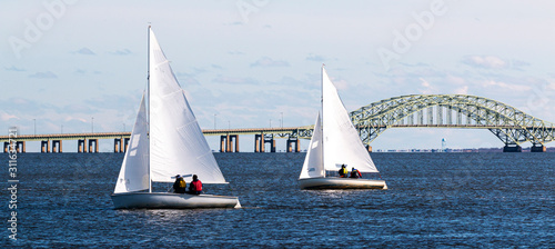 Two two person sailboats sailing toward West Islip with the Great South Bay bridge
