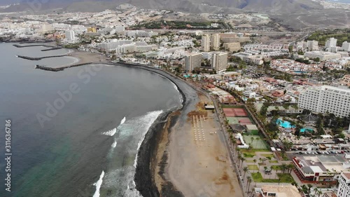 Aerial view of Las Americas city, Tenerife, Canary islands. Cinematic 4K footage with black volcanic rocky coast, Atlantic ocean, waves, city modern villas, quay with people, mountains. Bird eye shot photo