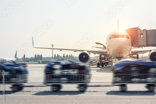 Luggage motion blurred trolley cart going fast delivering passenger baggage to modern plane on taxiway at airport on bright sunny day. Commercial aircraft on background at sunset or sunrise time