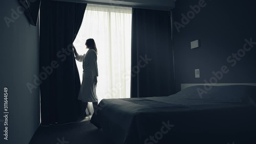 Rear view at young woman in bathrobe opening curtains in bedroom enjoying the sunlight. Good morning. Feeling happy photo