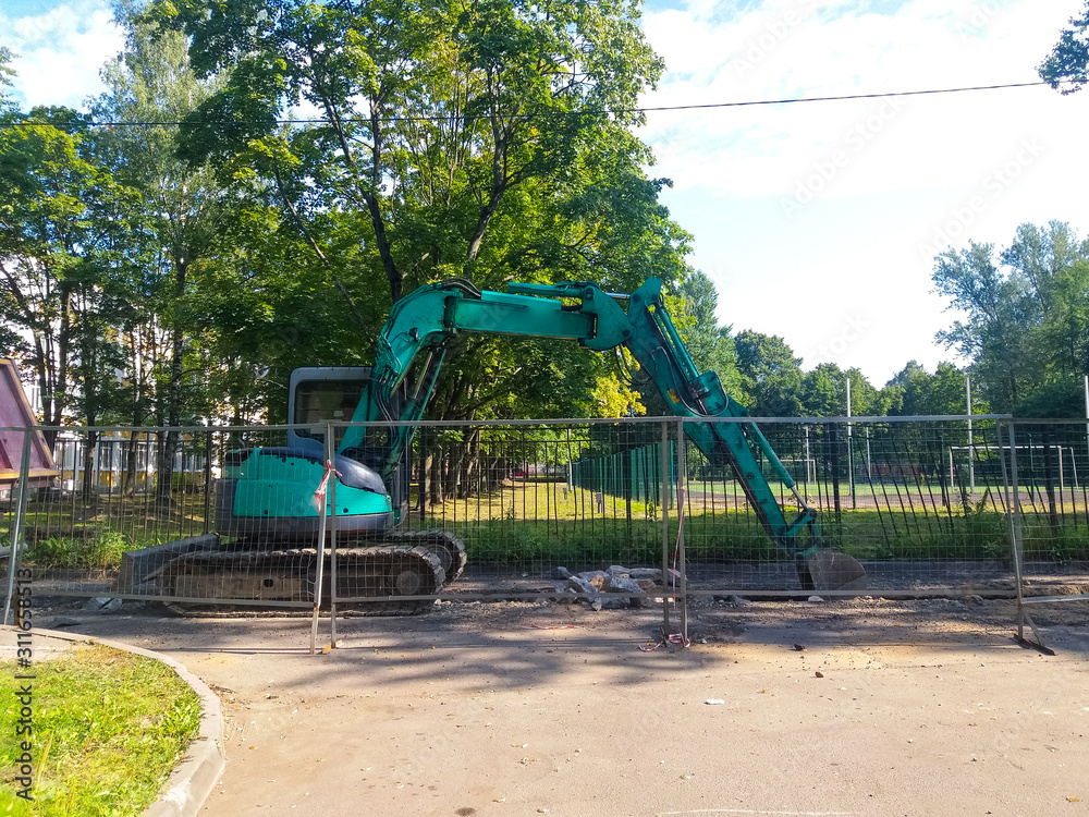 In the city yard, against the background of grass and trees behind a metal fence, a turquoise excavator digs up the roadbed. Russia, the city of St. Petersburg, August 2019. In the city yard, against the background of grass and trees behind a metal fence, a turquoise excavator digs up the roadbed. Russia, the city of St. Petersburg, August 2019.