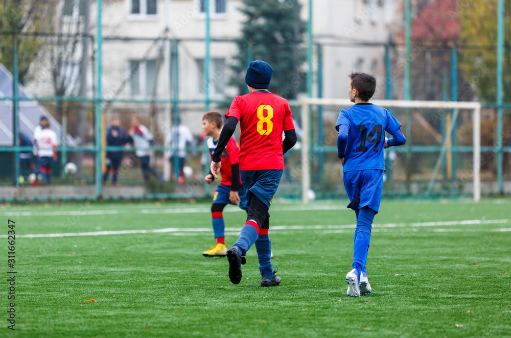 Obraz premium Boys in red and blue sportswear plays football on field, dribbles ball. Young soccer players with ball on green grass. Training, football, active lifestyle for kids concept