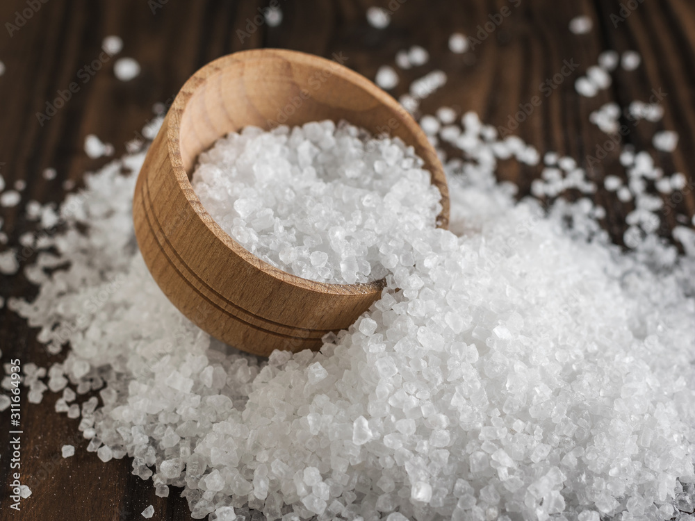 A wooden bowl on a pile of scattered sea salt on a wooden table. A wooden bowl on a pile of scattered sea salt on a wooden table.