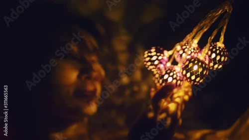 Magic hot light from lanterns falls on the woman eyes. The naked adult sexy caucasian girl lies at night on the bed and holds warm lamps in her hands. Christmas toy in black background. Indoor. photo