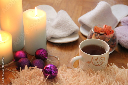 burning white candles stand on the floor  next to lies a fluffy plaid  two pairs of slippers and a mug of tea