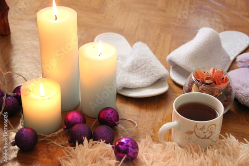 burning white candles stand on the floor, next to lies a fluffy plaid, two pairs of slippers and a mug of tea