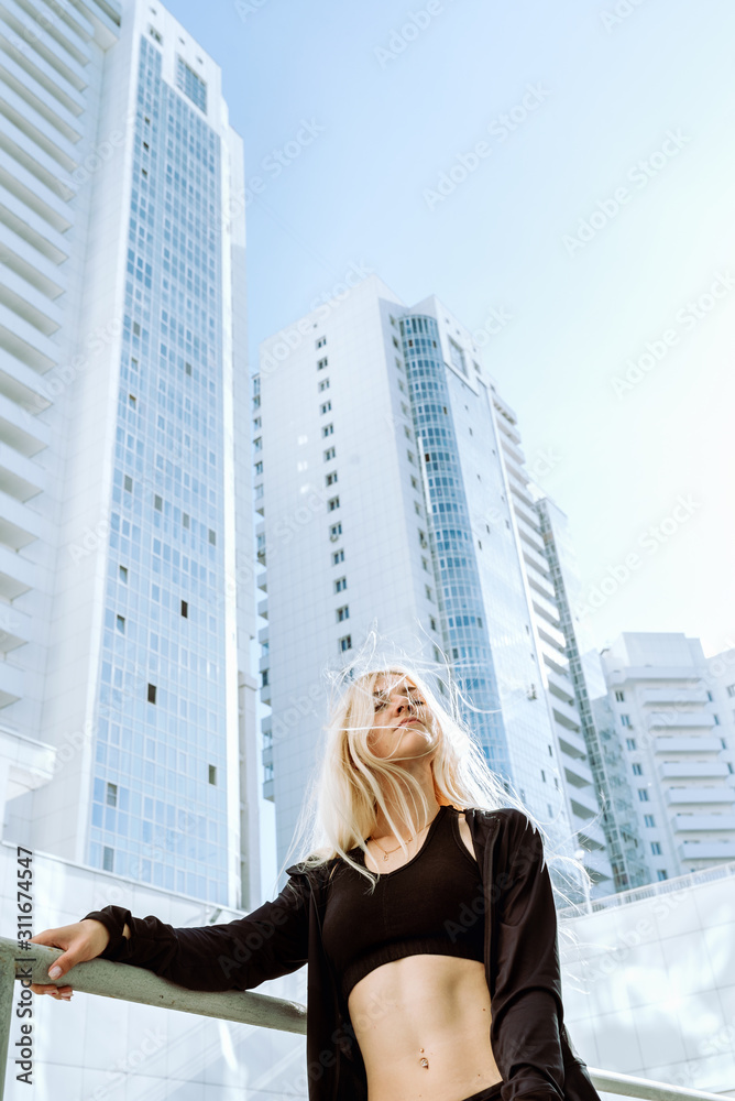 Young adult blond female athlete in black sport clothes standing in front of tall city buildings, selective focus