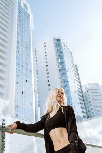 Young adult blond female athlete in black sport clothes standing in front of tall city buildings, selective focus