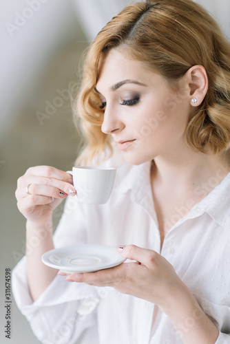 Beautiful young blonde woman  in a light shirt in a bright room enjoys a morning cup of coffee before going to work.Girl in the bathroom in candid clothes.