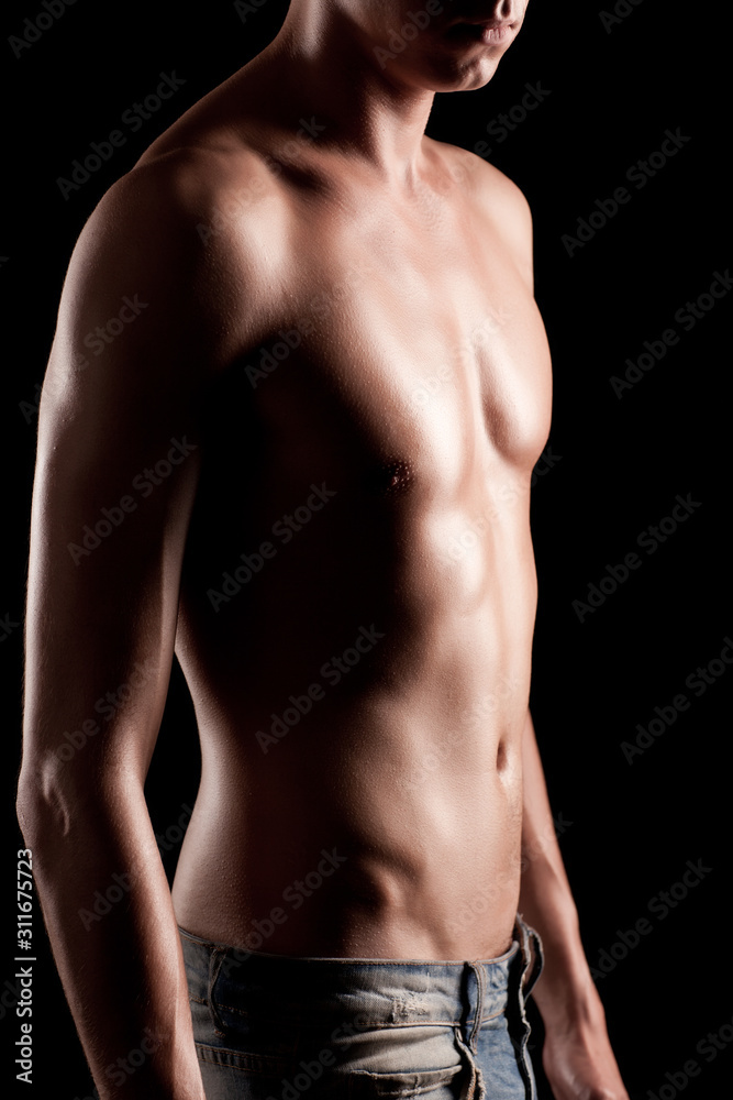 Young athletic man with naked torso in jeans standing over black background in photo studio. Beauty of man body and body relief concept