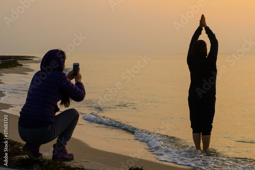 Salute to the Sun concept. Silhouette of people at sunrise. Girl photographs a woman who welcomes the rising sunon the seashore.  photo