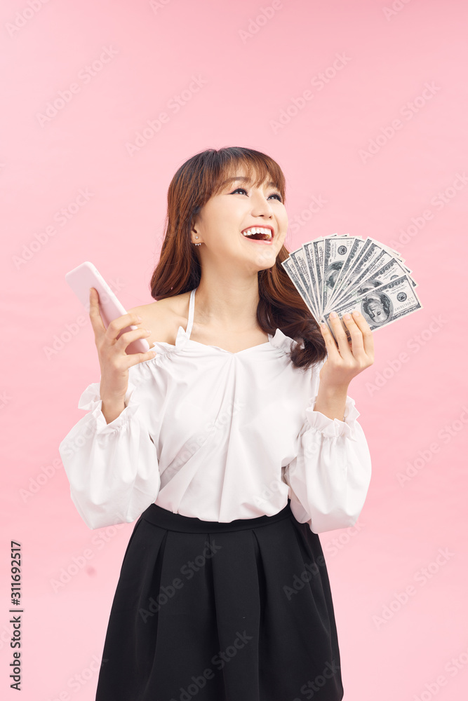 Portrait of a smiling young girl in dress standing over pink background, holding mobile phone, showing money banknotes Portrait of a smiling young girl in dress standing over pink background, holding mobile phone, showing money banknotes