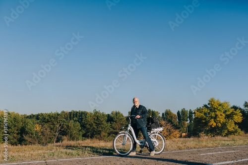 An elderly man  a pensioner rides an electric white bike on an asphalt road against the background of autumn nature. The concept of a happy old age. Fatigue and rest on the sidelines.