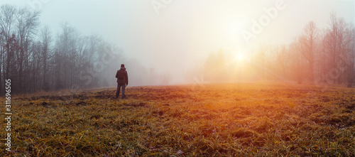 Panoramic view young man standing in jakcket and pants with backpack on meadow and trees with sun