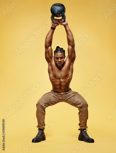 Strong young man swinging a kettlebell. Photo of handsome man with naked torso on yellow background. Strength and motivation. Full length