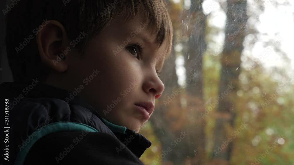 Five years old boy looking through dirty window of the train on green trees. Travel concept. Young traveler waiting for train stop. Child expression of sadness on the face of the boy. Side view
