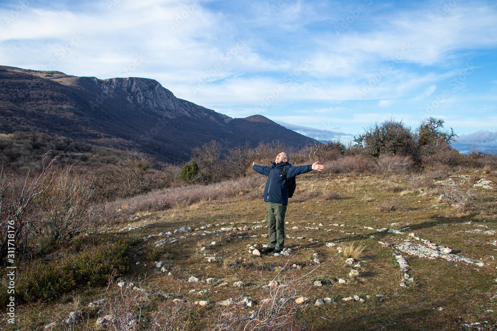 A smiling happy adult man, a hiker, with backpack, with his arms spread wide, stands in the center of a spiral laid out of stones, on top of Paraguilmen Mountain. Esoteric place of power. Lifestyle.