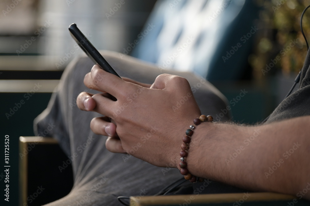 A young spanish man with trendy jewellery at his arm and fingers sits on a wooden chair while he is writing messages on his smartphone.