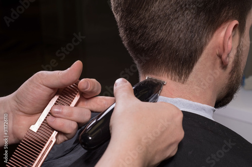 Hairdresser cuts hair with a machine for a young guy with a beard. Close-up of a master hand cutting a mans hair