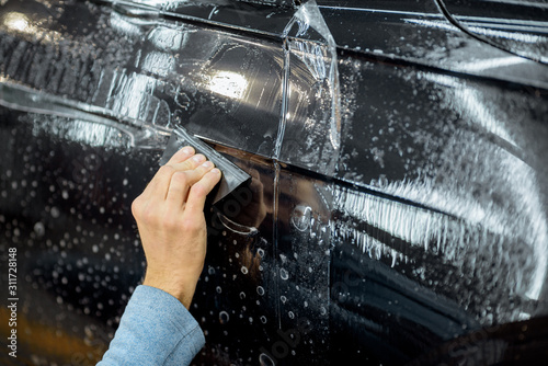 Fototapeta Naklejka Na Ścianę i Meble -  Worker sticking anti-gravel film on a car body with scrapper at the detailing vehicle workshop, close-up. Concept of car body protection with special films