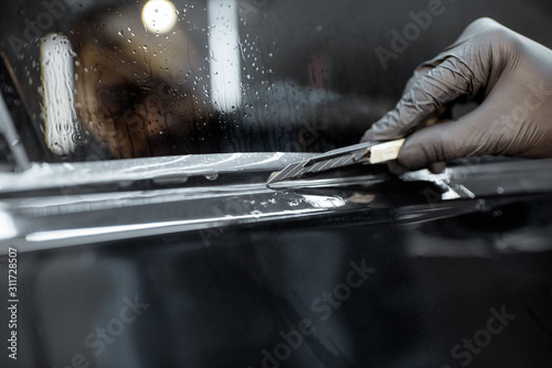 Worker trimming with cutter remains of a protective film, sticking it on a car body at the vehicle service, close-up. Concept of car body protection with special films