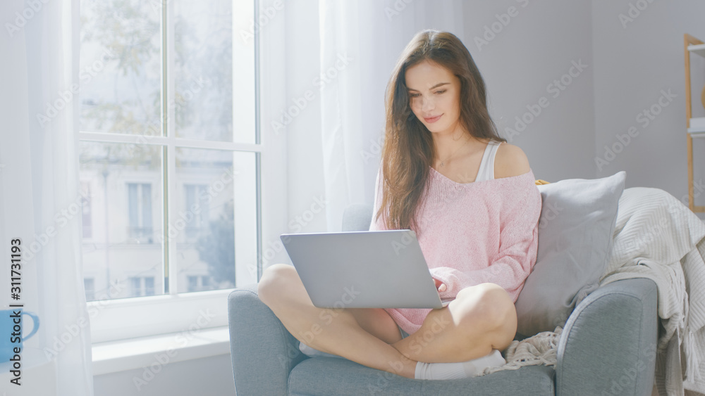 Portrait of Young Brunette Works on Laptop Computer at Home in a Cozy Living Room. Gorgeous Girl Wearing Pink Sweater, Spends Relaxing Time. Portrait of Young Brunette Works on Laptop Computer at Home in a Cozy Living Room. Gorgeous Girl Wearing Pink Sweater, Spends Relaxing Time.