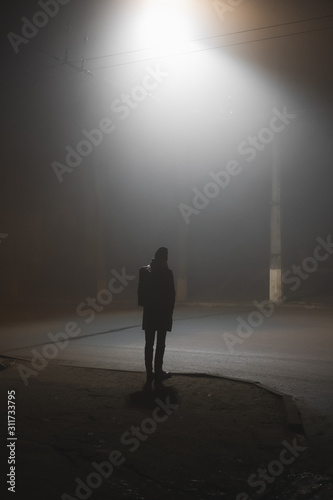 A person standing near the road in a foggy town at night. Noir aesthetics  concept of loneliness  dullness and late autumn mood