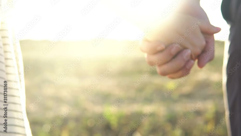 Man and woman holding hands and slowly walk away through the field. Man takes hand of woman in white dress and then they go while the sun shines brightly showing their silhouettes.