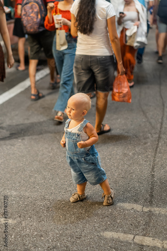 Small child in a stylish denim overalls enjoys a walk in the evening among passers-by.