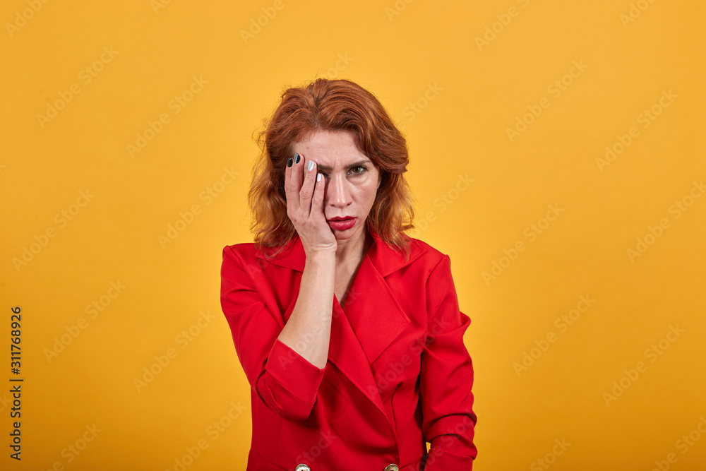 Disappointed caucasian young woman covering eye with hand, looking at camera wearing fashion red jacket over isolated orange background. People lifestyle concept.