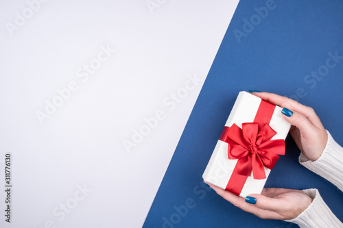Gift box with a red bow on a gray and blue background in women's hands. Space for text.