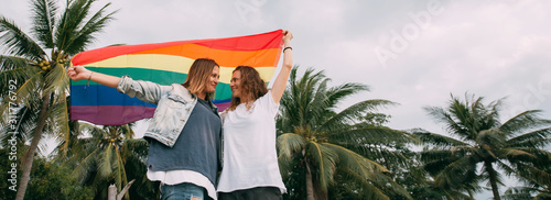 Fotografie, Tablou Two women with rainbow flag on the beach on a background of palm trees