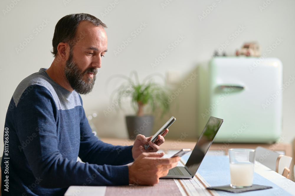 side view of young bearded man with a smartphone in each hand and his laptop on the table Multitasking, work interruptions side view of young bearded man with a smartphone in each hand and his laptop on the table Multitasking, work interruptions