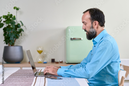 side view of young man in blue shirt working at home with his laptop. Simple but modern decoration with soft colours