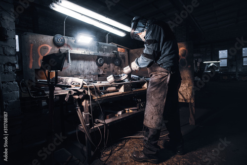 At the dark workshop experienced worker in protective uniform is working with metal.
