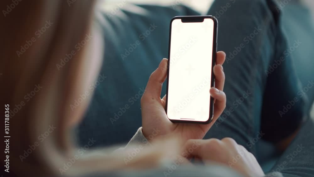 Over shoulder shot of woman holding a mobile phone or smartphone with a vertical green screen in her hand at home lying on the sofa. Girl tap on display and watching content.