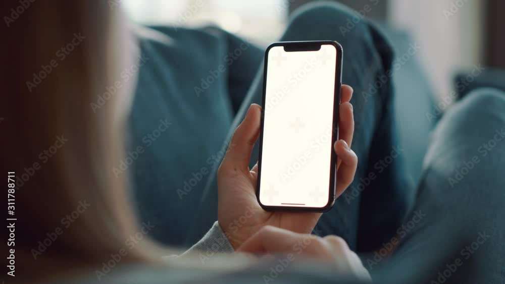 Close up view of young woman with blonde hair lying on blue couch and tapping on green screen in light modern room. Girl using smartphone or mobile phone, wathing video, content, blogs. Slow motion.