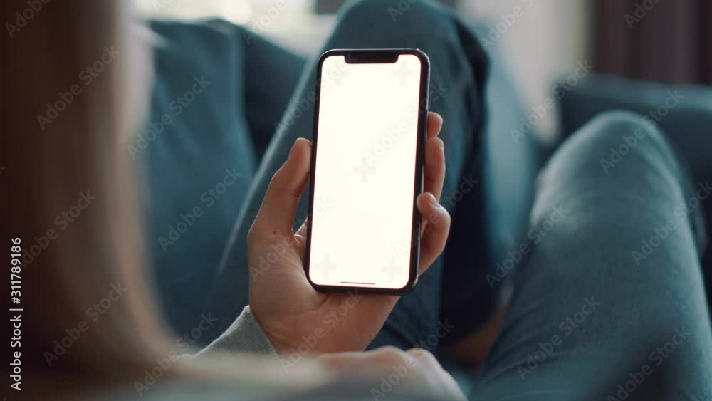Girl with blond hair lying on blue couch and holding smartphone with green screen in her hand at light modern room. Relaxed woman looking at the mobile phone.