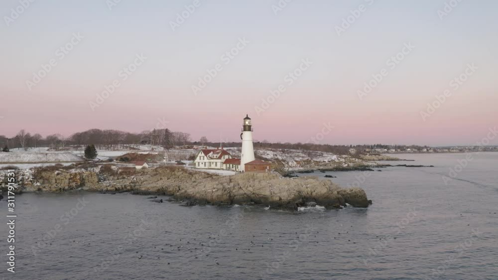 Lighthouse on rocky coastal outcrop above floating sea ducks, winter sunrise, SLOW AERIAL ORBIT Lighthouse on rocky coastal outcrop above floating sea ducks, winter sunrise, SLOW AERIAL ORBIT