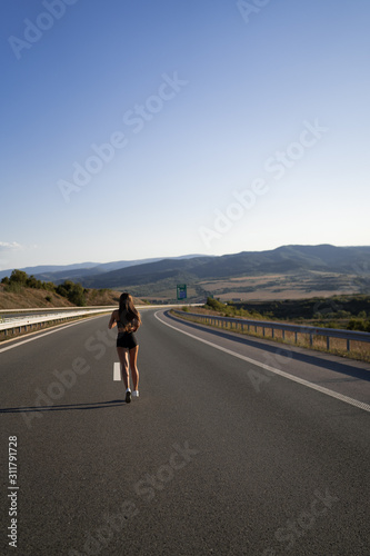 Young girl running along the road. Athletic happy woman jogging in trendy black sexy top and shorts enjoying the sun exercising. Healthy lifestyle. Perfect fitness body shapes and tan skin