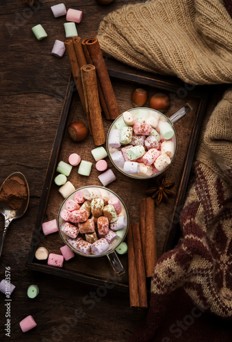 Hot winter or autumn drink with cocoa, white chocolate, spices and colorful marshmallows in cups on vintage wooden background with knitted warm sweater, top view with copy space