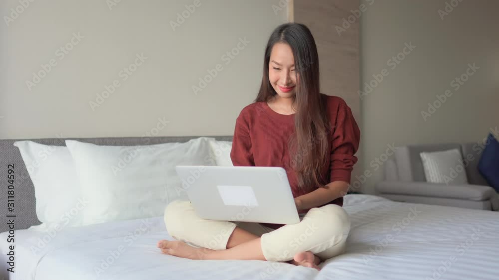 Happy Asian Female Sitting on Bed and Typing on Laptop. Working From Home Concept With Copy Space Happy Asian Female Sitting on Bed and Typing on Laptop. Working From Home Concept With Copy Space