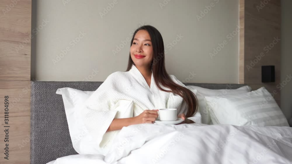 Asian woman with long dark hair sitting in bed wearing robe holding cup and saucer. Young attractive woman smiling and relaxing in bed with cup of tea or coffee. Bright sunlight on happy face. Asian woman with long dark hair sitting in bed wearing robe holding cup and saucer. Young attractive woman smiling and relaxing in bed with cup of tea or coffee. Bright sunlight on happy face.