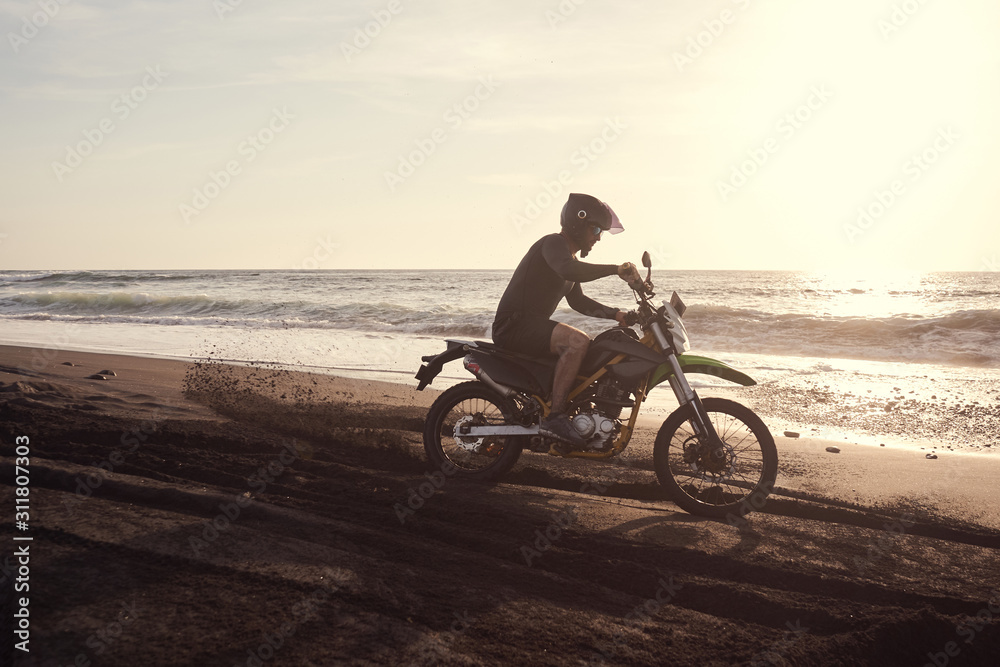 Fototapeta premium Male masculine model with cross road motorbike at the ocean coast with black sand at the golden hour time