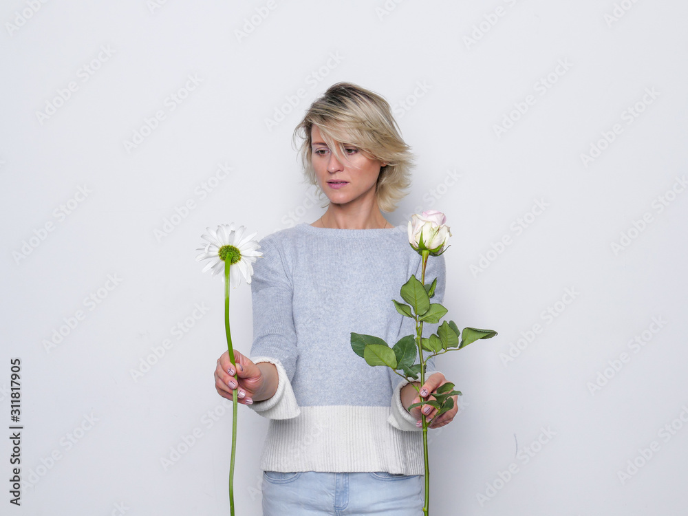 Portrait of young blonde beautiful woman with makeup in natural casual dress choosing between two flowers Portrait of young blonde beautiful woman with makeup in natural casual dress choosing between two flowers