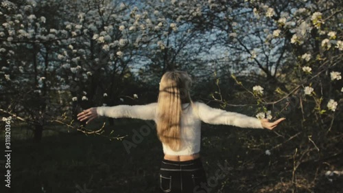 Attractive young woman in sweater, jeans is dance and spinning among blossom apple tree. Sunset, spring orchard and green field. Slow motion. Caucasian one girl, long fluttering hair. Sun glasses. photo