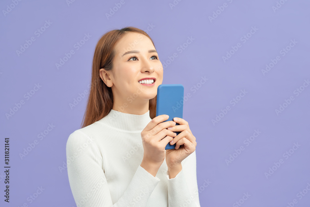 Smiling asian woman holding smartphone and looking away over violet background Smiling asian woman holding smartphone and looking away over violet background