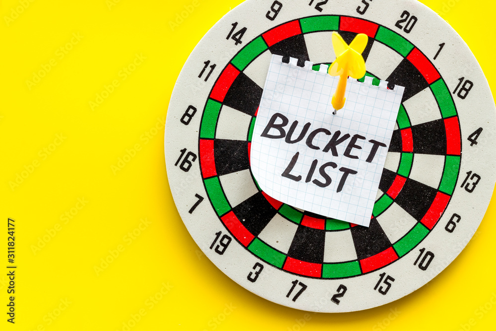Bucket list attached by dart to dart board on yellow background top-down copy space Bucket list attached by dart to dart board on yellow background top-down copy space