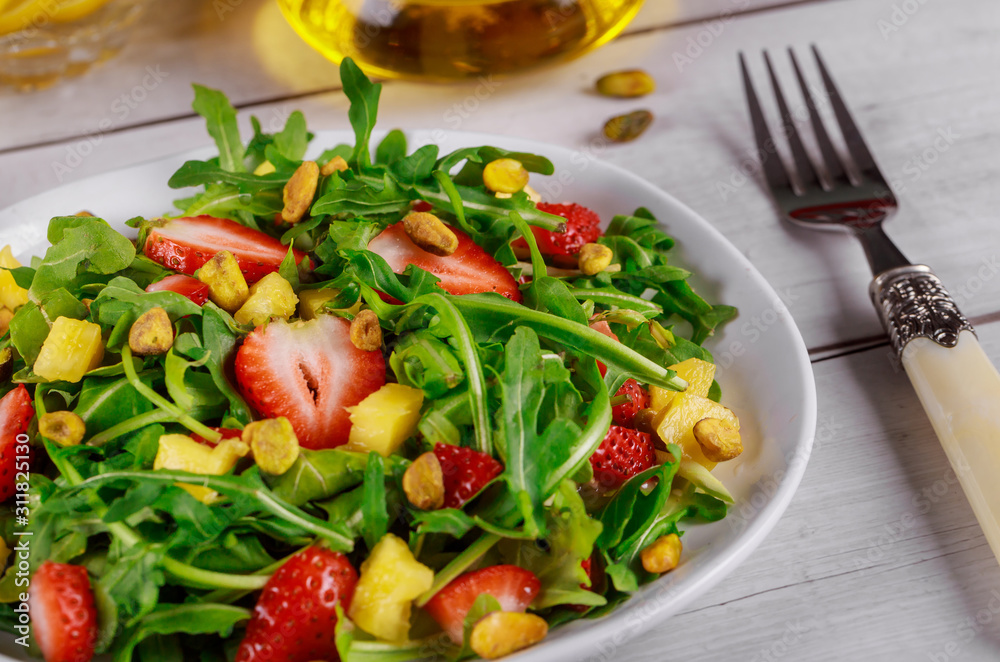 Healthy fresh salad with arugula, strawberries, pineapple and pistachios served on white plate on rustic wooden table.